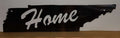 Black metal sign shaped like the state of Tennessee with 'Home' engraved on it, placed on a wooden surface against a white wall.
