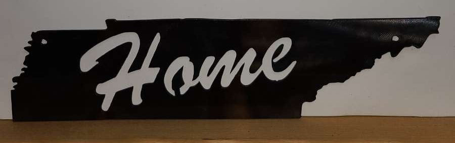Black metal sign shaped like the state of Tennessee with 'Home' engraved on it, placed on a wooden surface against a white wall.